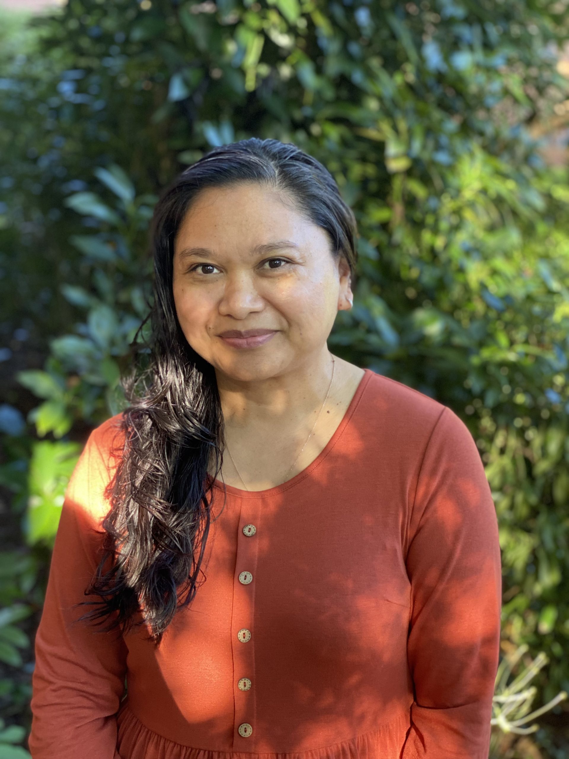 Ms. Maria is smiling at the camera. She has long brown hair and is standing in front of vibrant greenery.